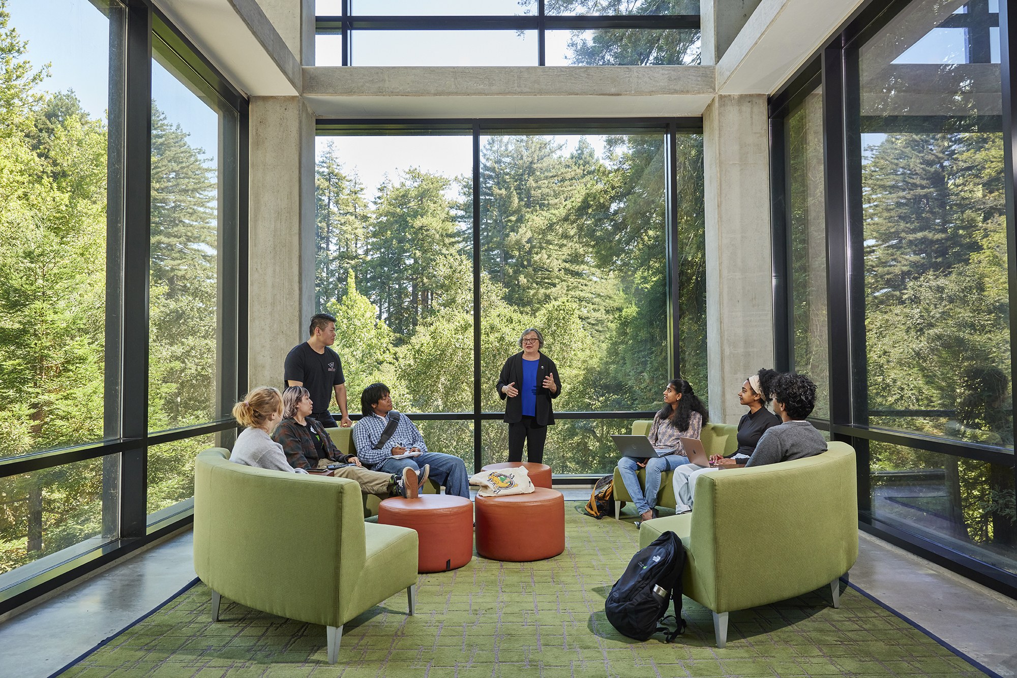 Chancellor Larive in the library talking with students in front of a big glass window with redwood trees in the background